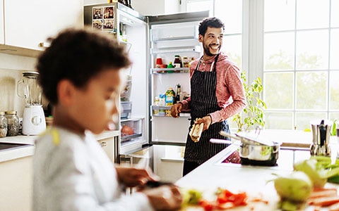 背景中的男子站在打开的冰箱旁边，前景中的一个孩子正在桌子上玩食物