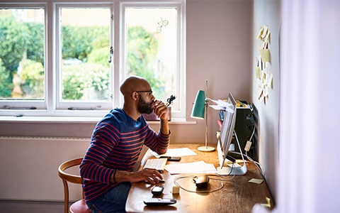 一名男子在家里工作时坐在窗前的办公桌前，为在家工作的员工提供网络安全服务