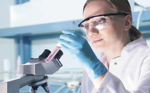 一位女士穿着实验服，戴着护目镜，在实验室里拿着试管，看着显微镜。